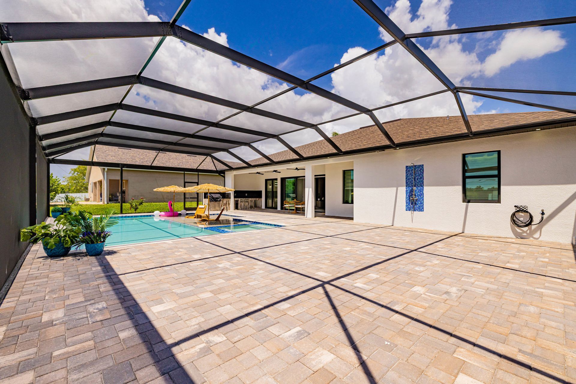 Patio with a pool, enclosed by a black screened frame. White house and blue sky in background.