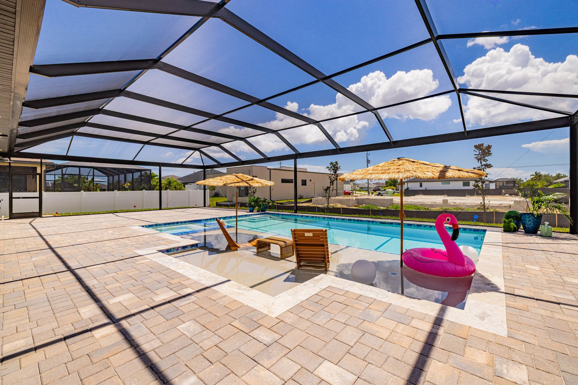 Pool area with screen enclosure; lounge chairs, flamingo float, umbrellas, and sunny blue sky.