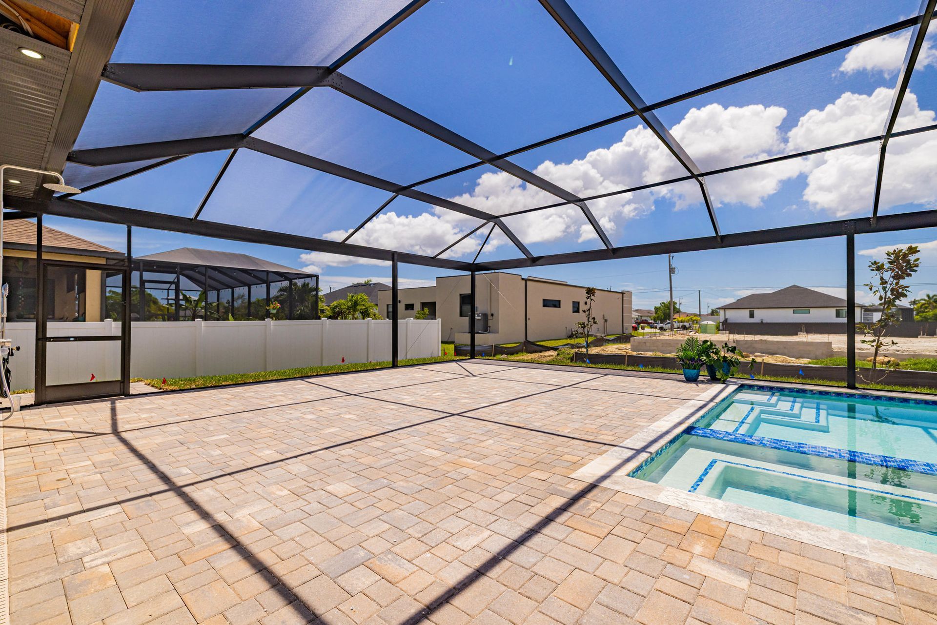 Screened-in patio with pool and spa under blue sky with clouds. Brick pavers, houses in background.
