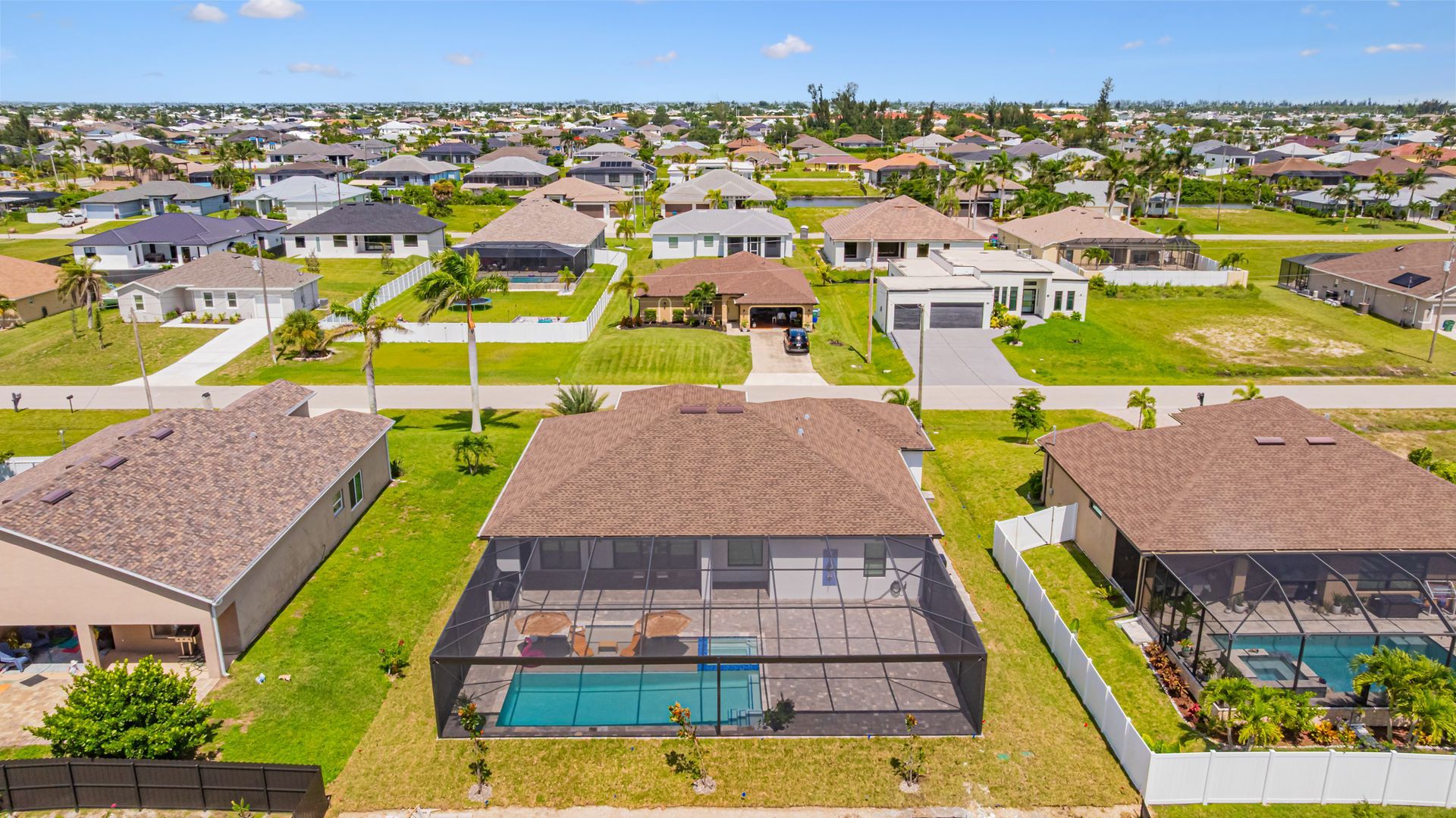 Aerial view of suburban homes with pools, lush lawns, and palm trees under a clear blue sky.