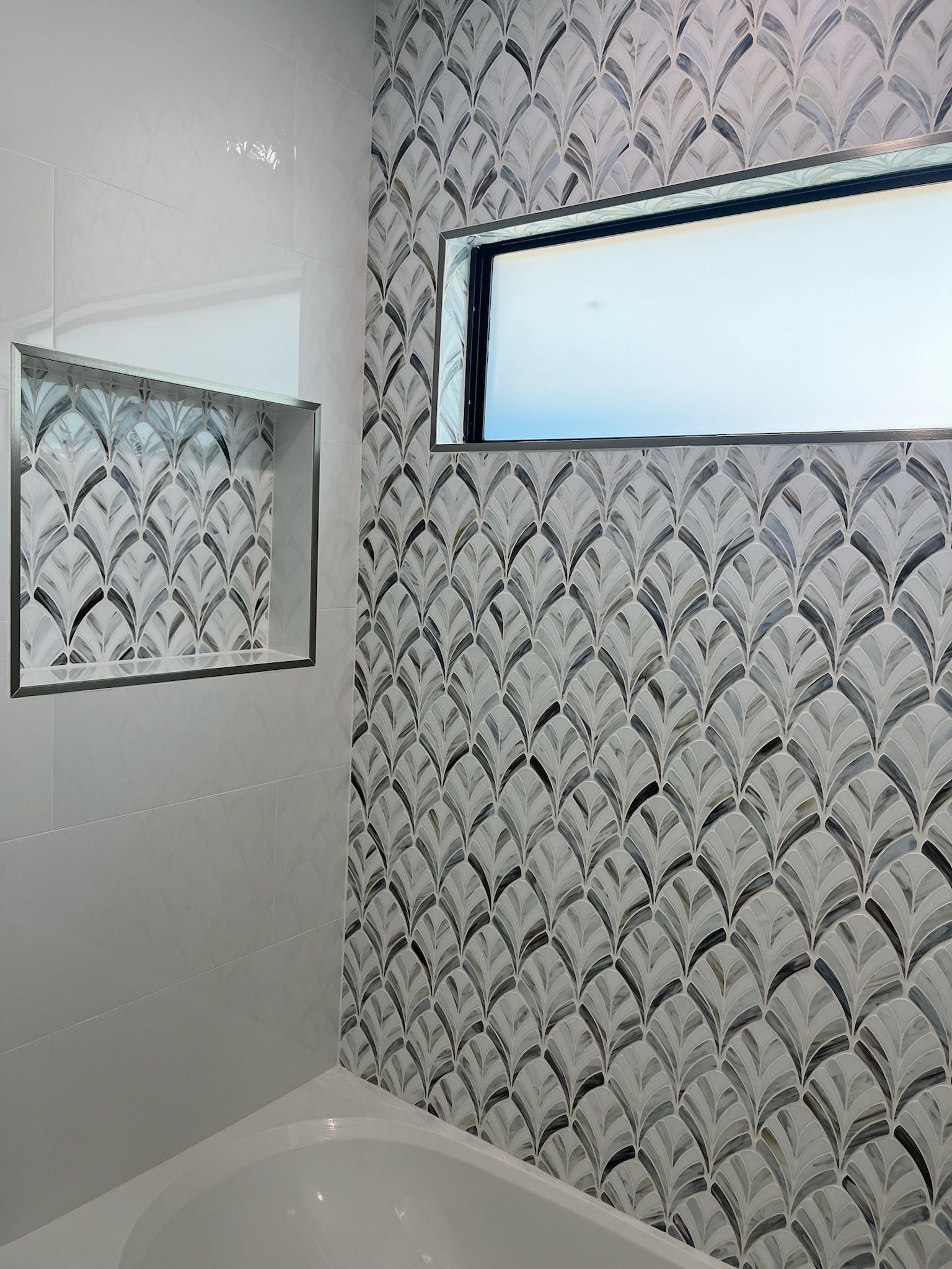 Bathroom with white tub and wall with niche and patterned mosaic tiles. Window over the tub.