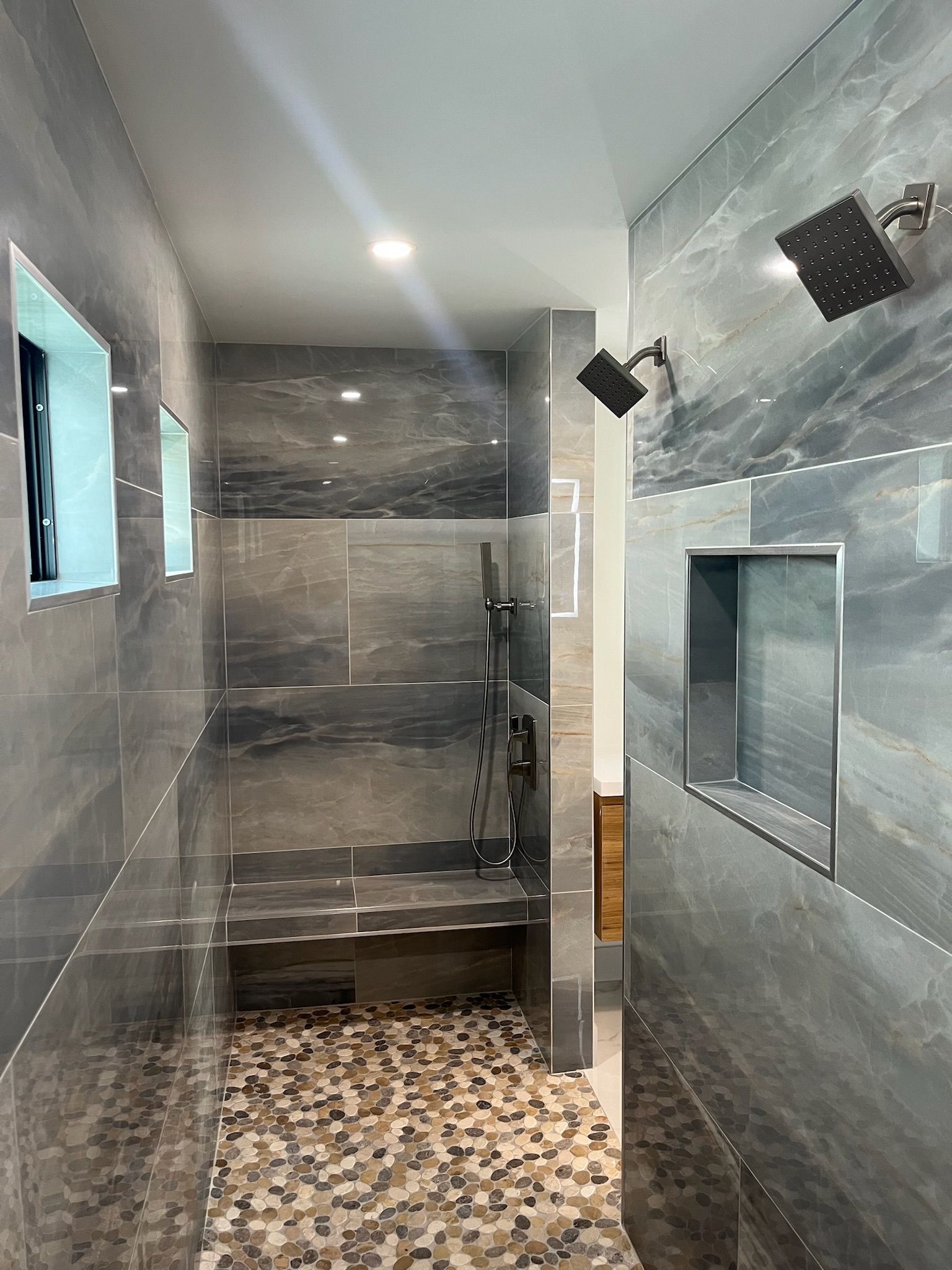 Spacious shower with gray tile walls, mosaic floor, built-in bench and shelves, dual shower heads.