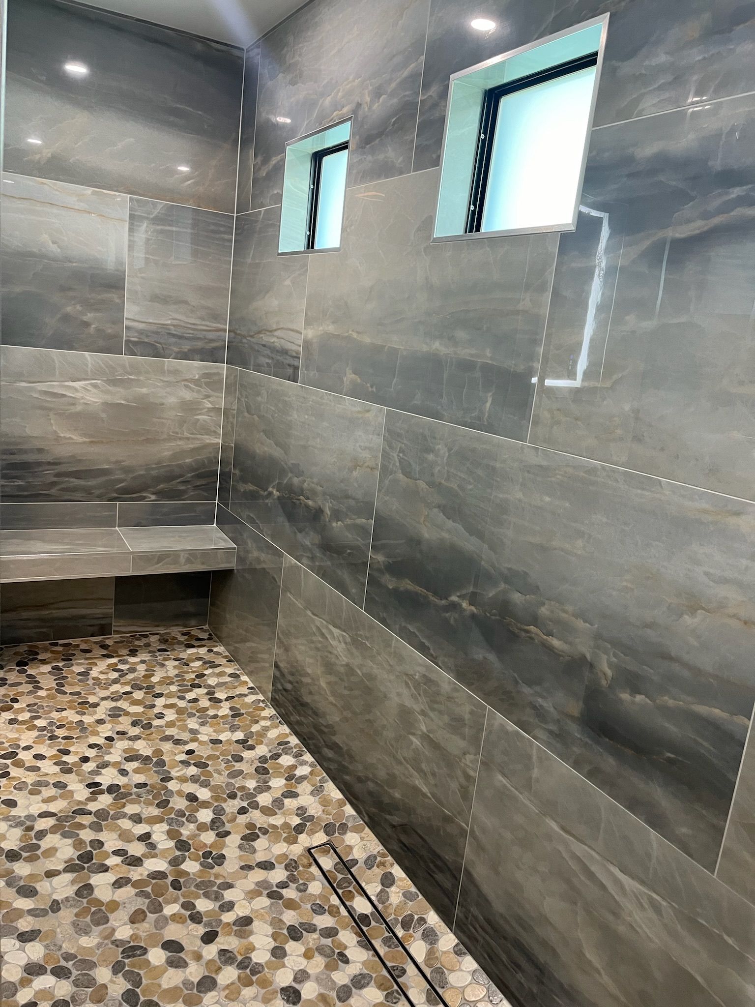 A stone shower with mosaic floor, built-in bench, gray textured walls, and two small windows.