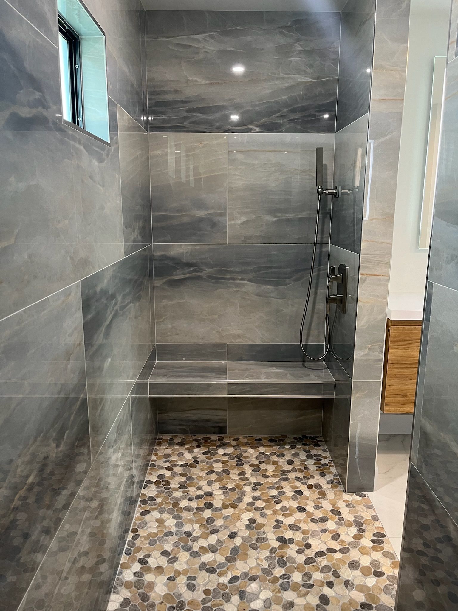Gray tile walk-in shower with mosaic floor, a built-in bench, and black shower fixtures.