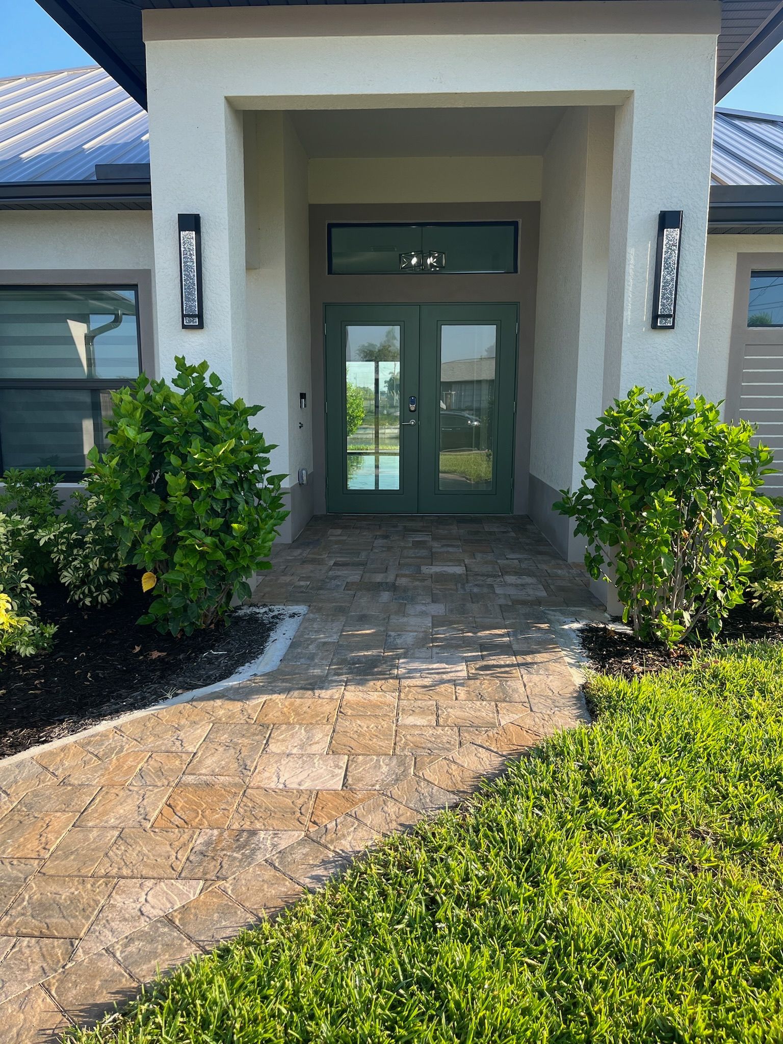 Brick path leading to green double doors of a house with bushes and black light fixtures.
