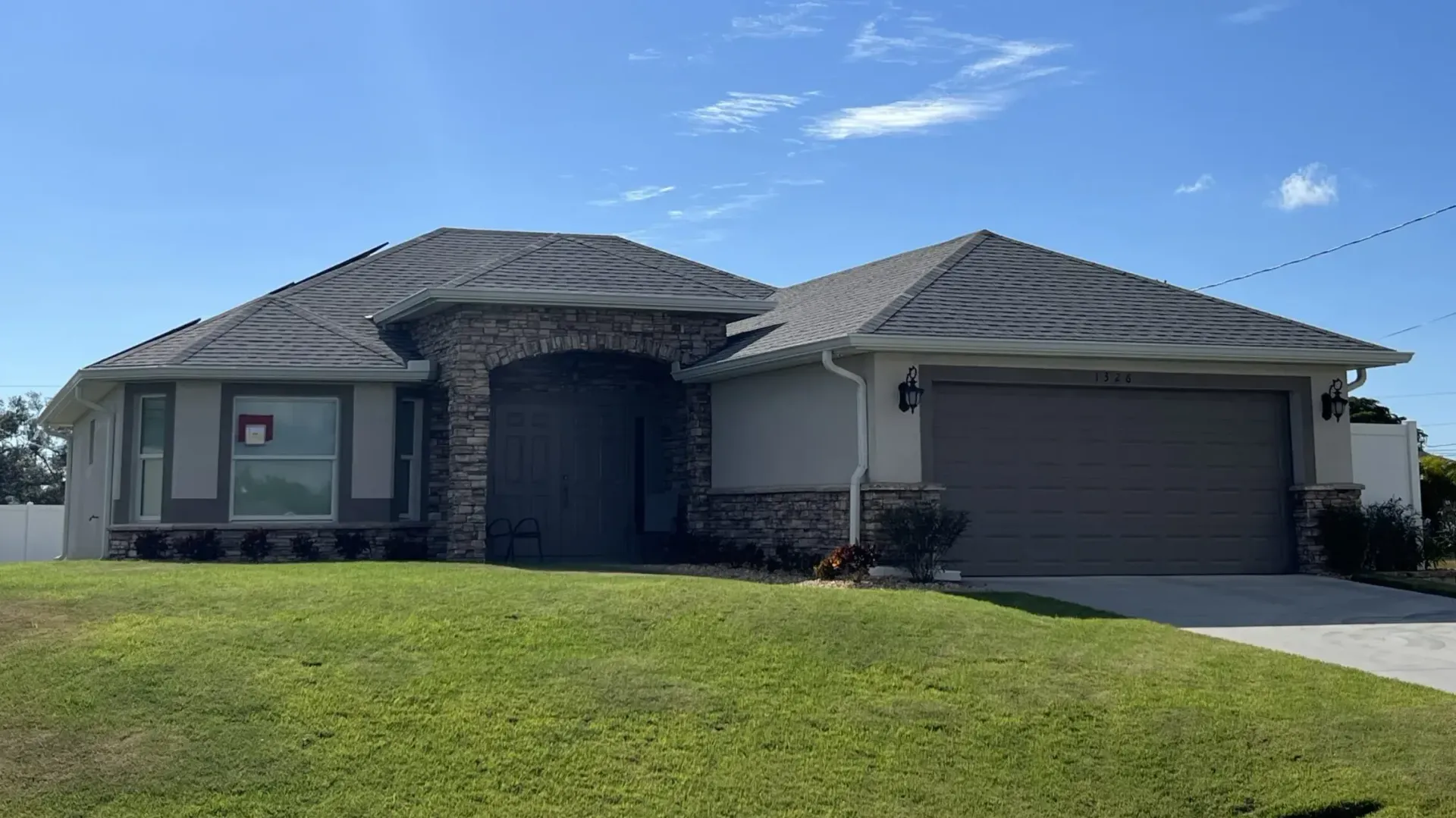 Single-story house with gray roof, stone facade, garage, and green lawn on a sunny day.