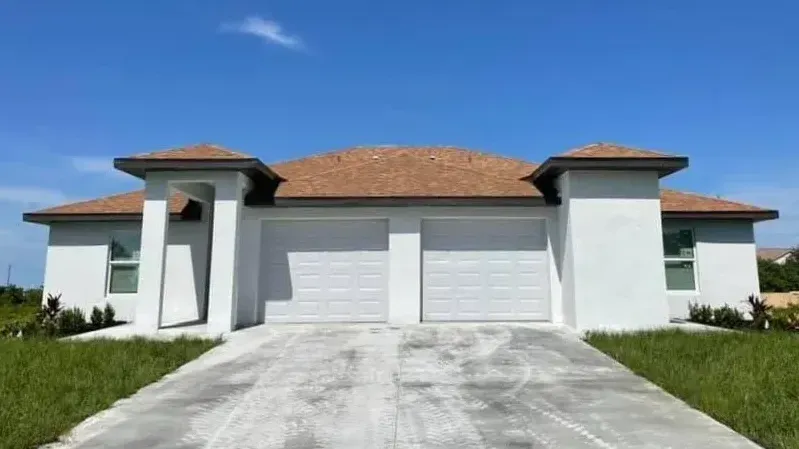 White duplex with two-car garage, brown roof, blue sky. Concrete driveway and small bushes in Florida.