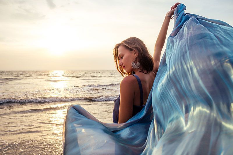 Virtual Dispensary -  Beautiful brunette girl in blue gray chameleon dress with long train sitting on a beach at sunset