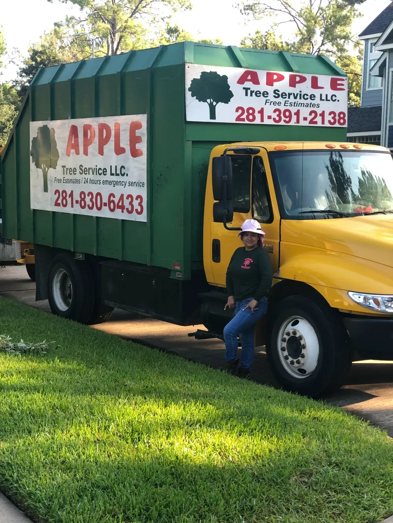 Woman in front of yellow Apple Tree Service truck. Green container, tree logo, pink ribbon, sunny day.