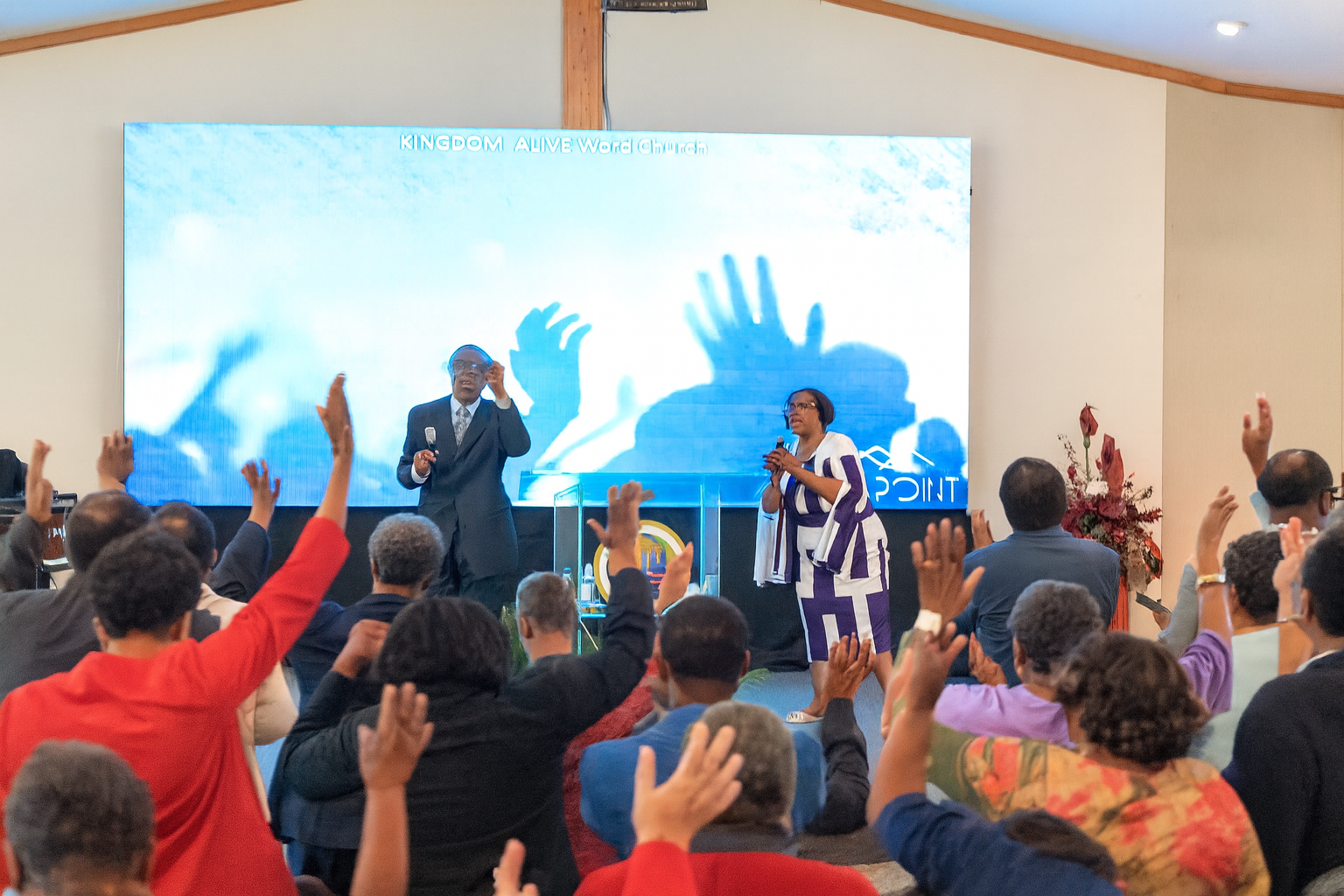 People with raised hands in church, two speakers, cross on wall.