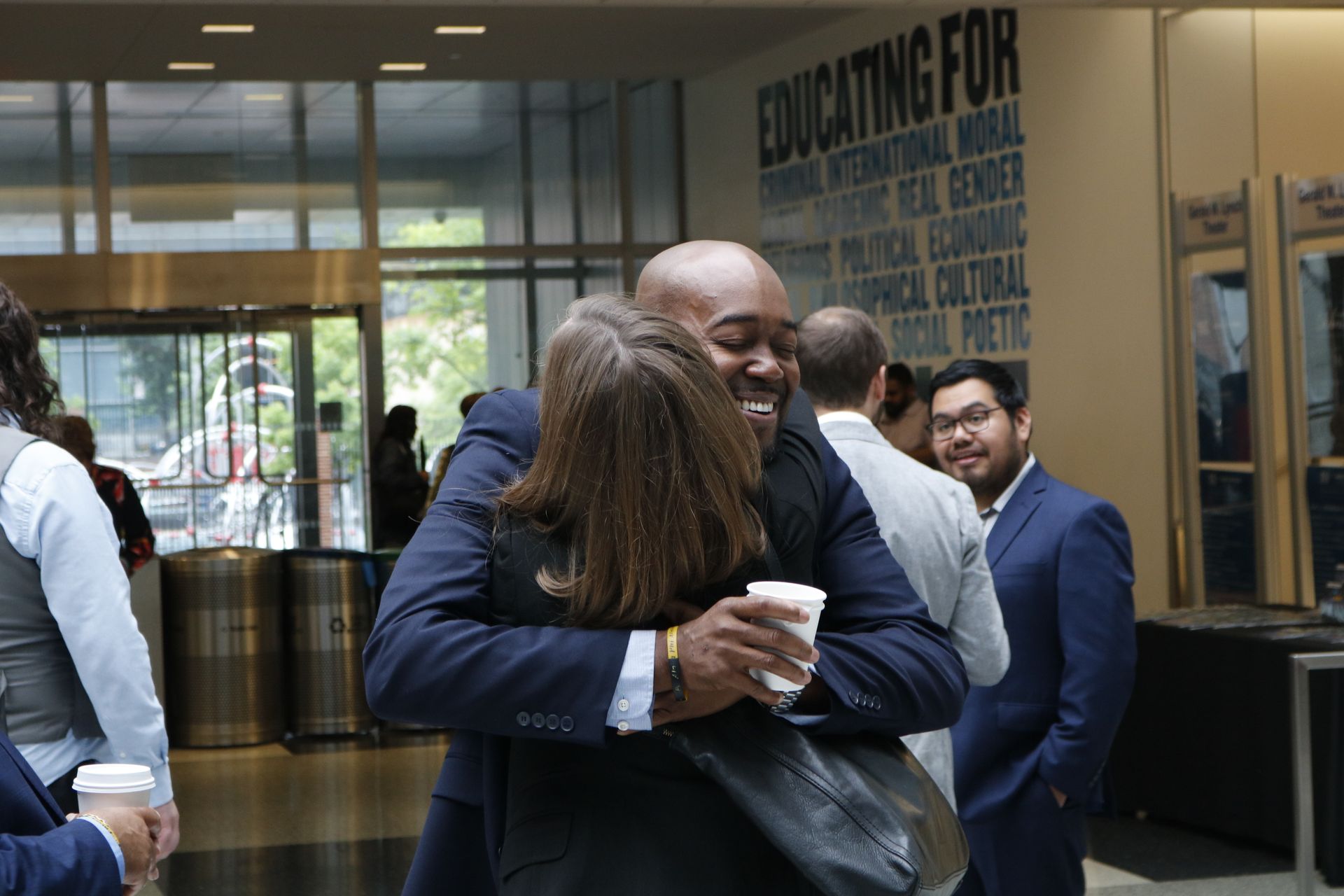 A man in a suit hugs a woman, smiling. Inside a building with others, holding coffee.