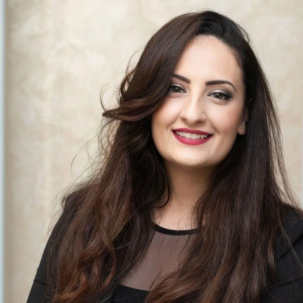 Donna con i capelli tirati indietro, che indossa un top nero, guarda direttamente la telecamera con un'espressione neutra.