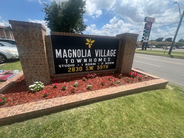 Magnolia Village Townhomes sign with brick accents, black background, and address 2830 SW 59th, with floral landscaping.