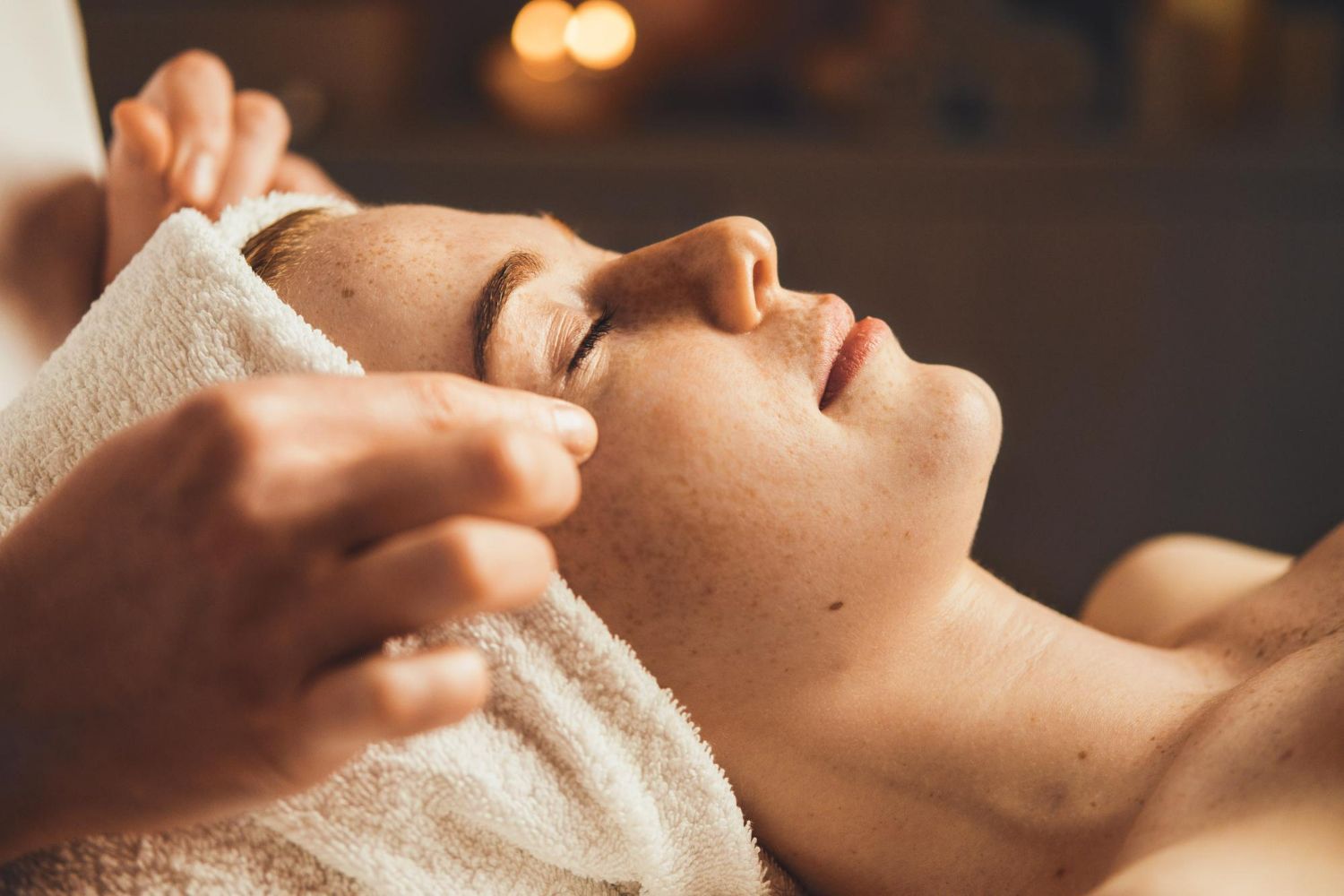 Person receiving a facial treatment; therapist's hands on forehead with a towel. Relaxed expression.