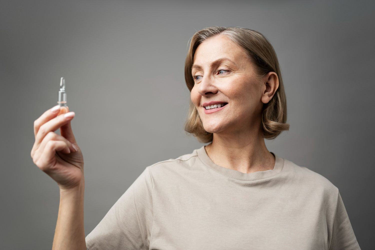 Woman holding and looking at a small glass vial, smiling. Gray background, beige shirt.