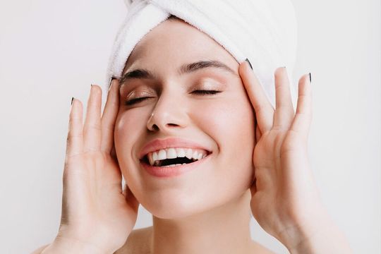 A woman with a towel wrapped around her head is smiling and touching her face.