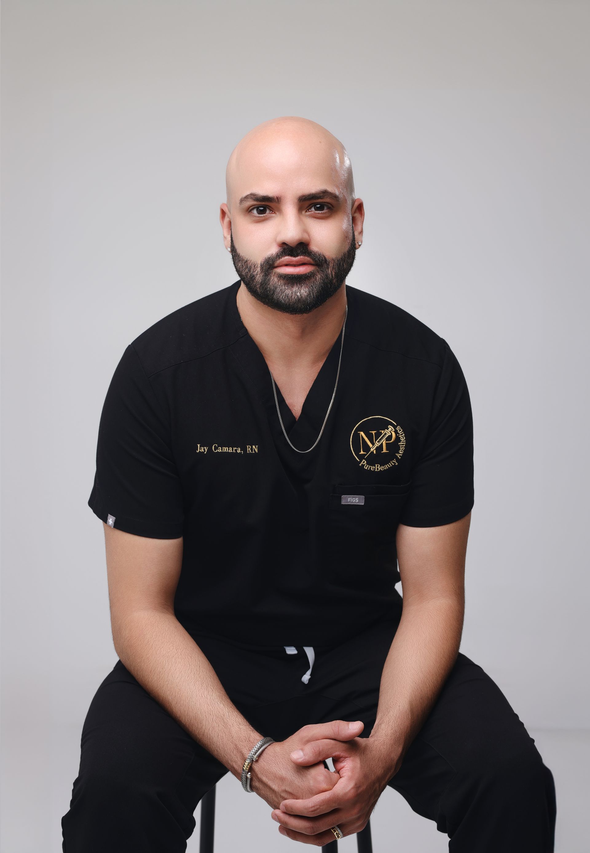 A bald man with a beard is sitting on a stool with his hands folded