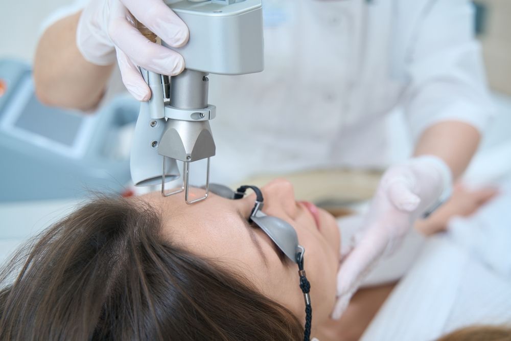Woman receiving laser treatment on her face; doctor in gloves holds device.