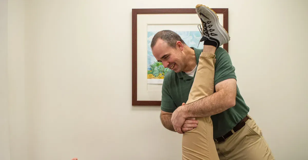 Man in green shirt stretching a person's leg up, smiling in a room with a framed picture.