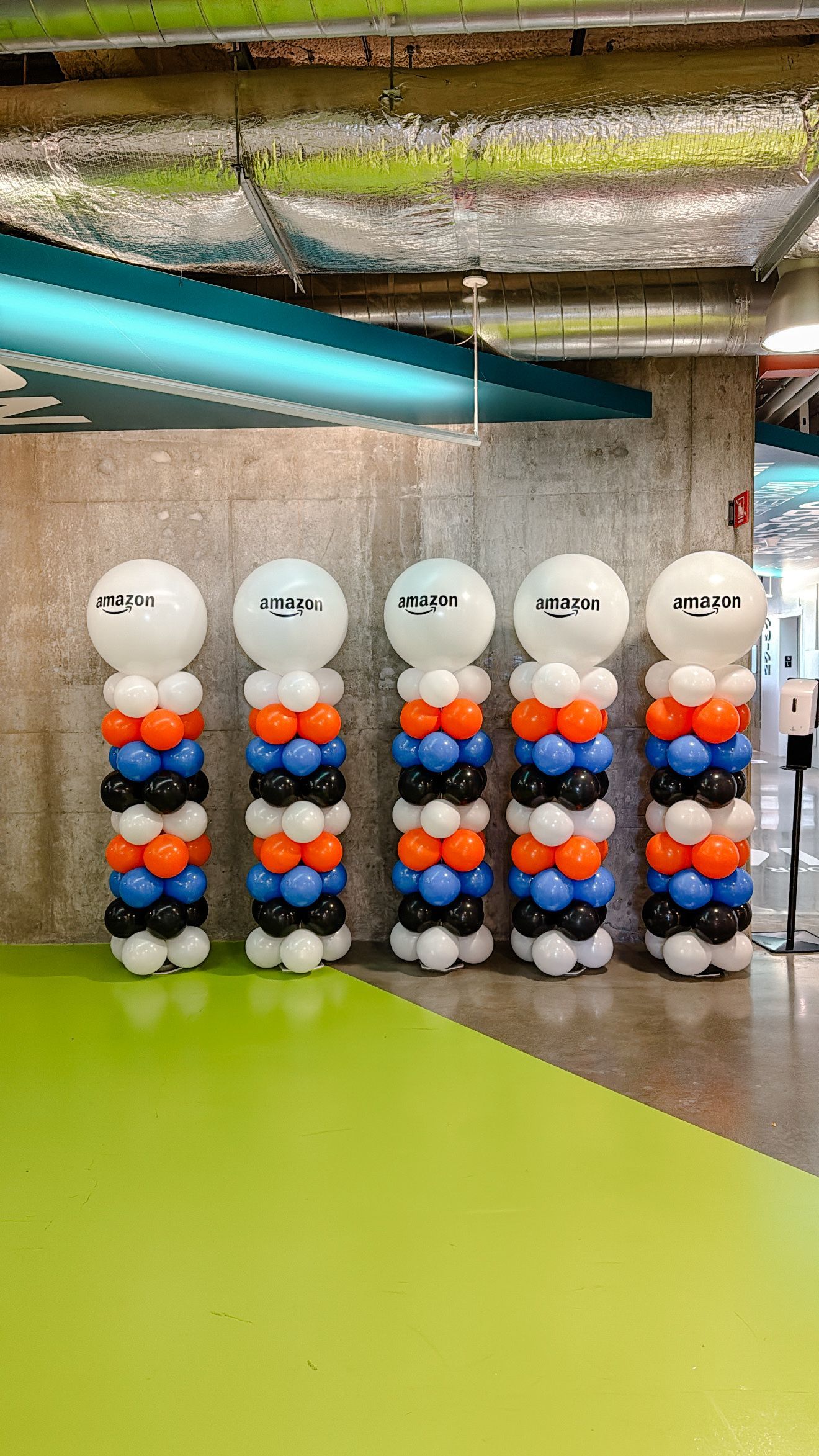 Five balloon columns in orange, blue, and black, topped with white balloons and text. In a room with green flooring.