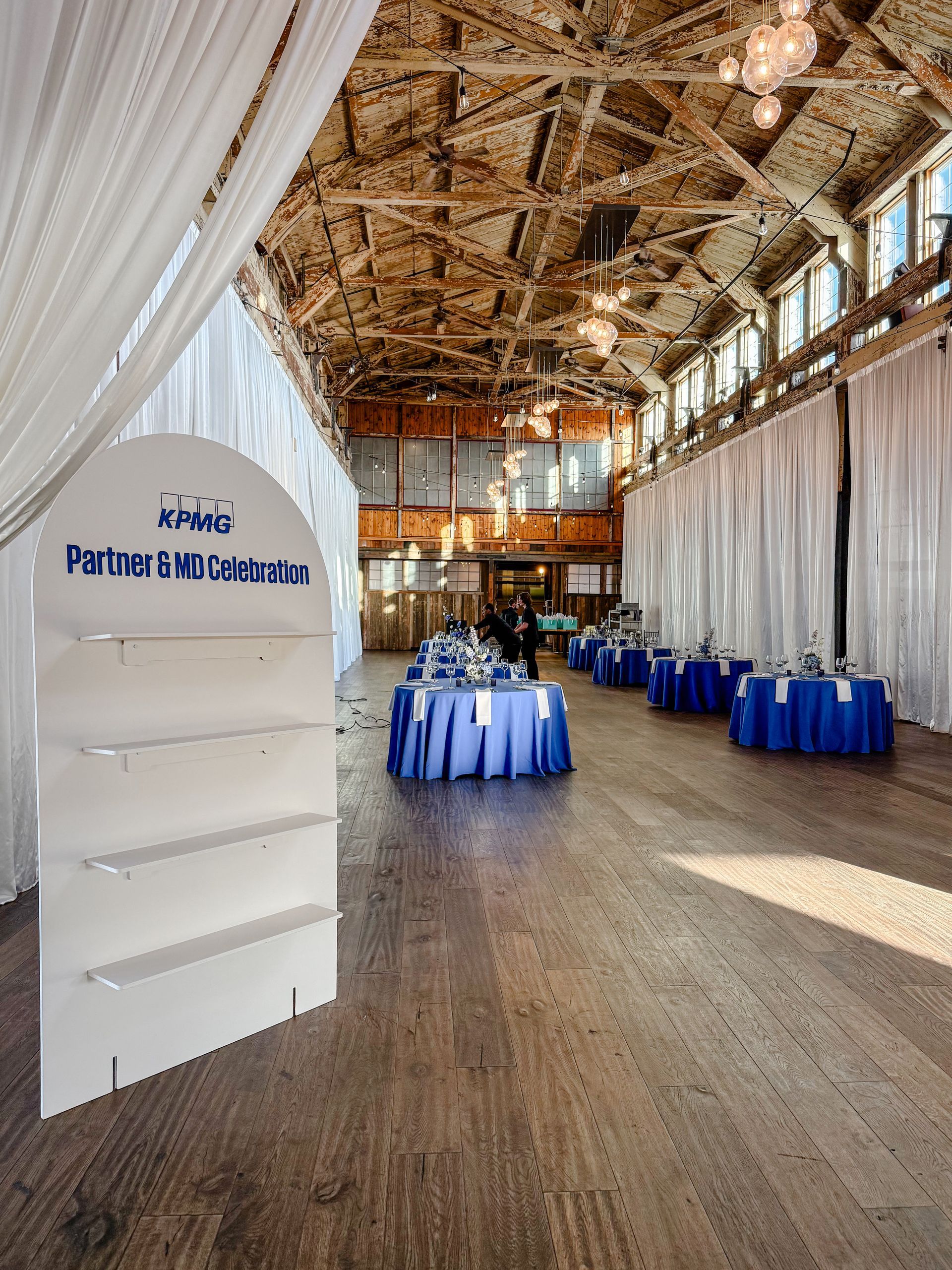 A display stand at the entrance of a rustic, high-ceilinged venue featuring draped white walls and tables with blue cloths.