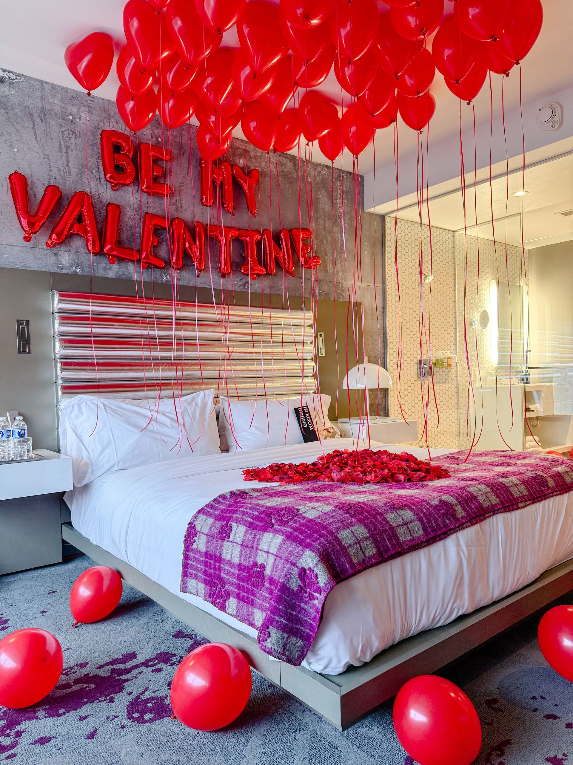 Romantic hotel room with red balloons, rose petals, and “Be My Valentine” decor over a white bed