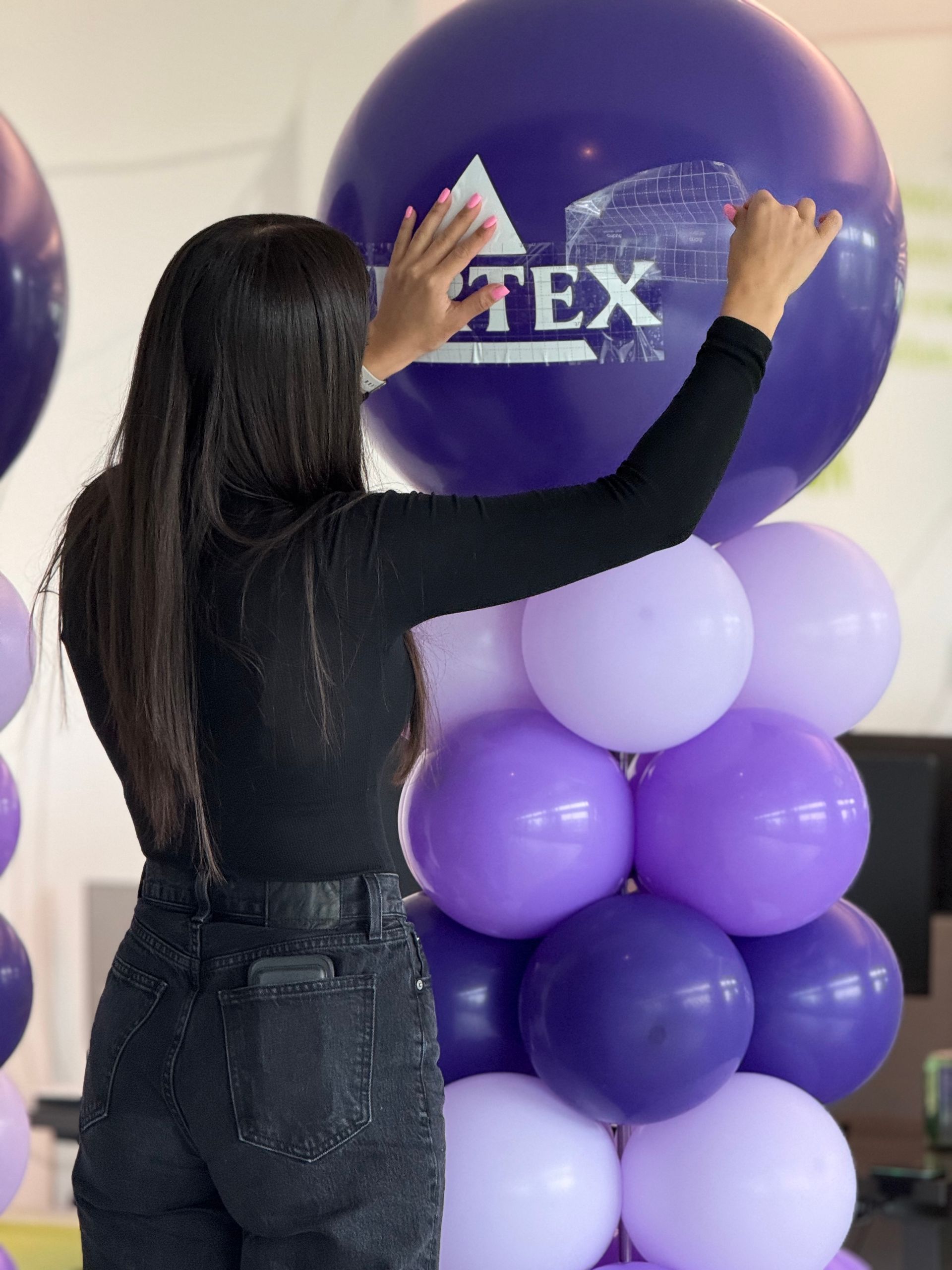 Woman arranging a large purple balloon with a logo on top of a cluster of smaller purple balloons.