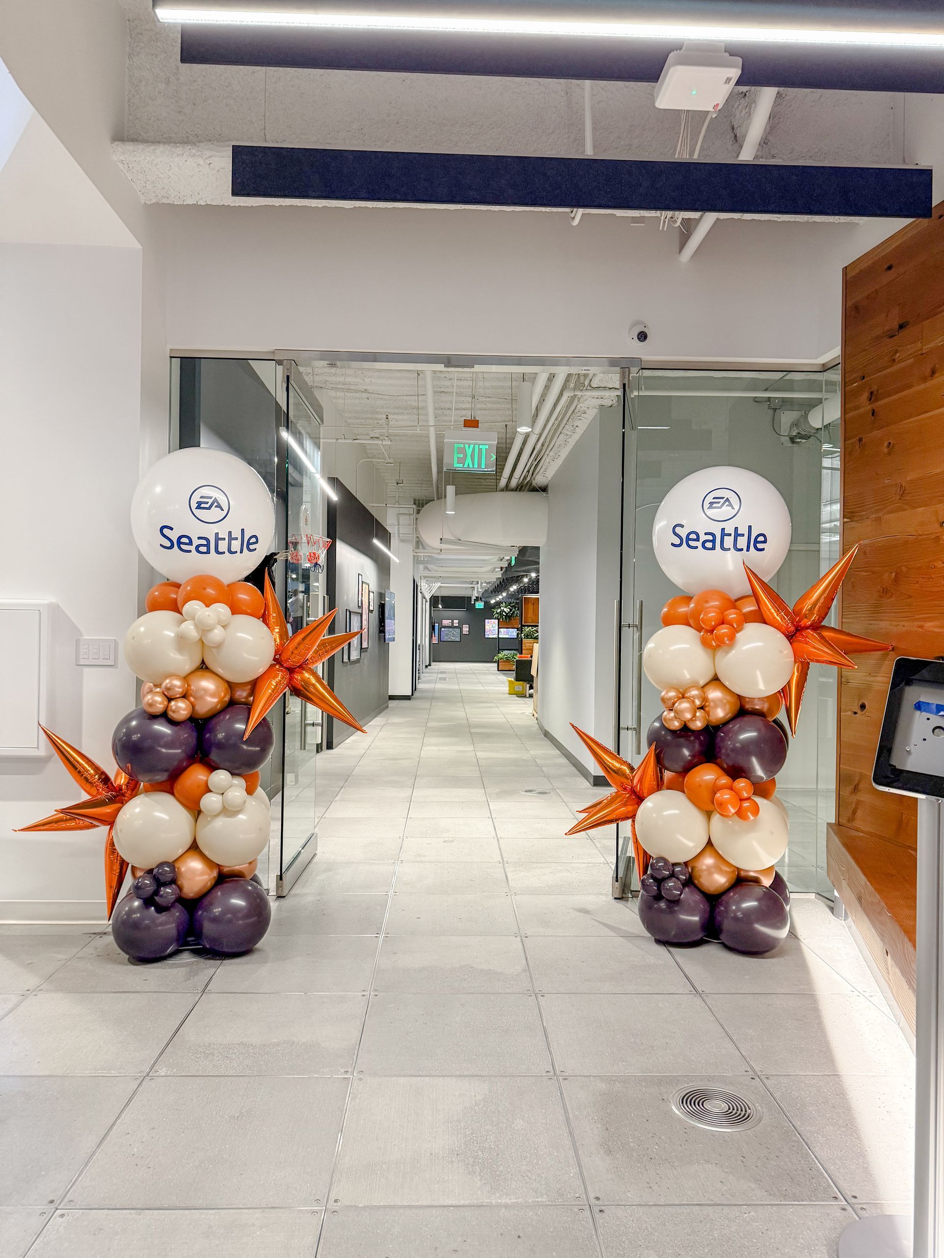 Two balloon columns mark a hallway entrance, with white, orange, and purple balloons, topped by logo balloons.