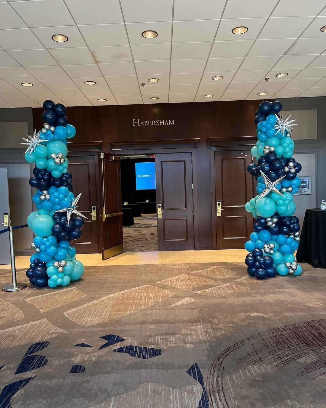 Two balloon columns, teal and navy, flank a doorway. The doorway reads