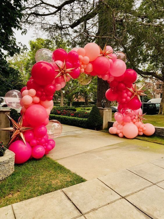 Balloon arch in shades of pink and red, adorned with gold stars, set outdoors.