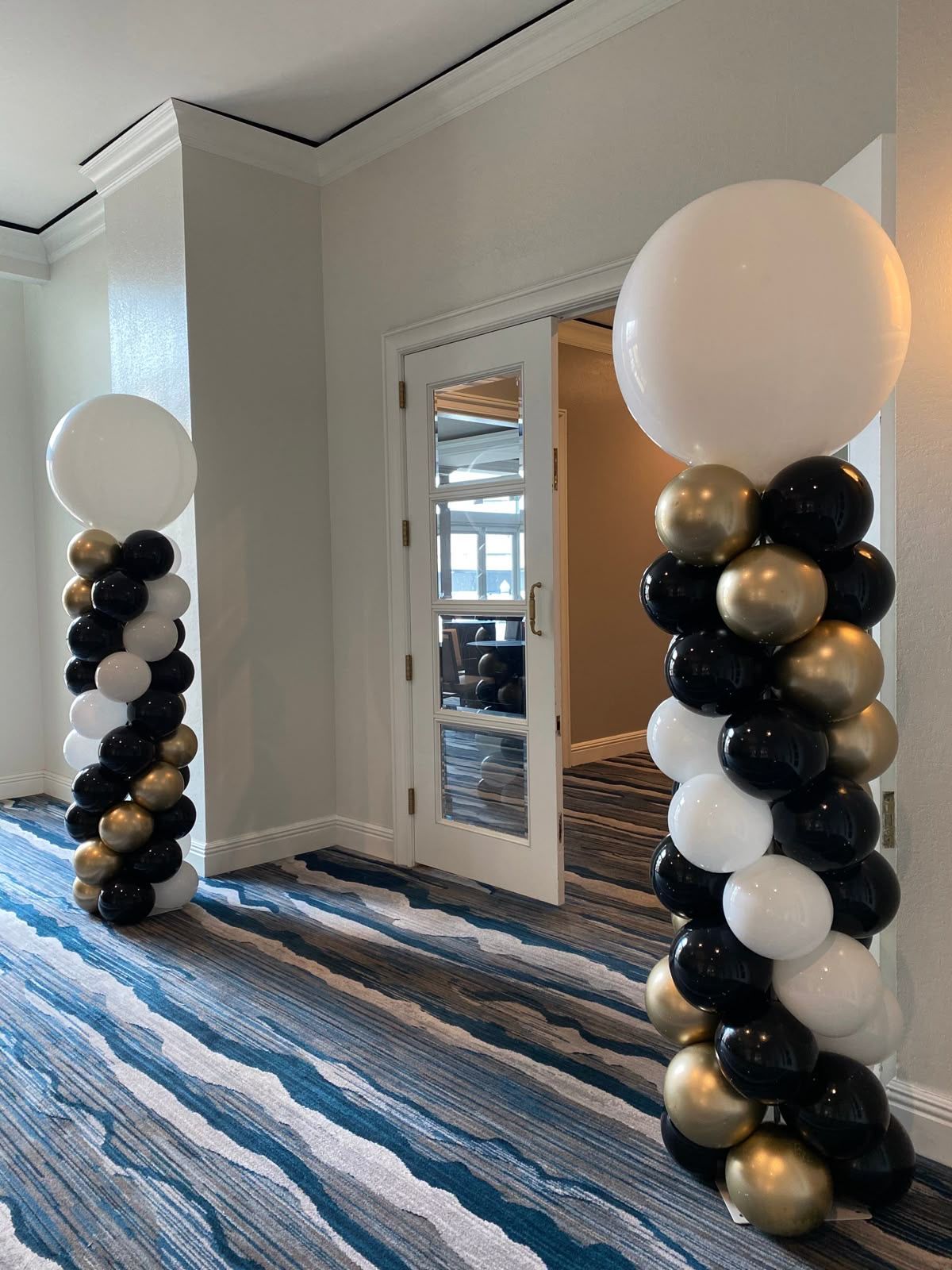 Two balloon columns, black, gold, and white, flank a doorway with patterned blue carpet.