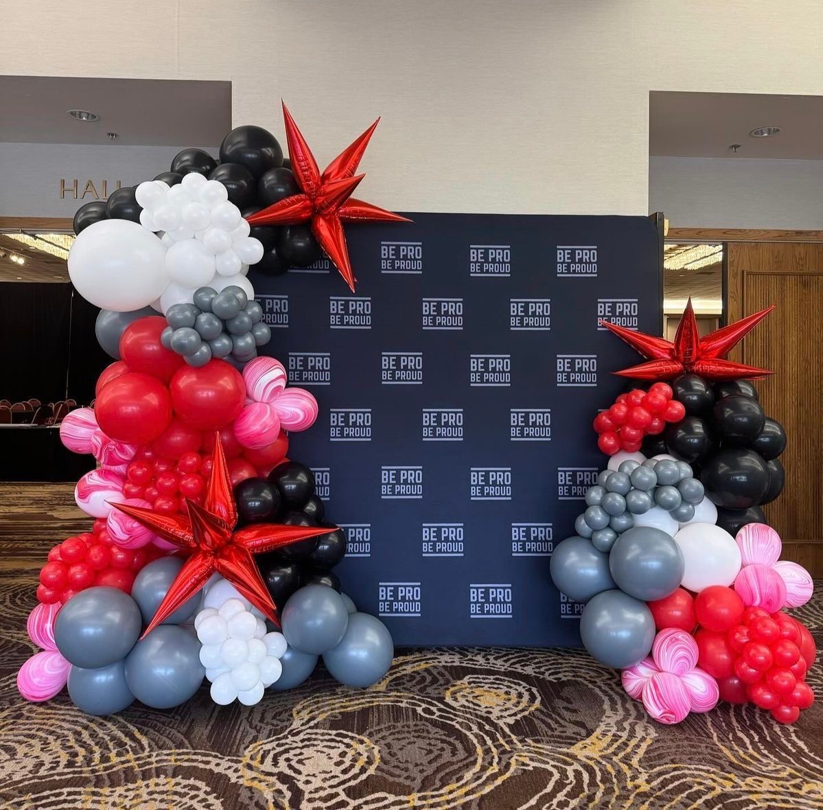 Event backdrop with balloons in red, black, gray, white, and pink, and red starburst accents.