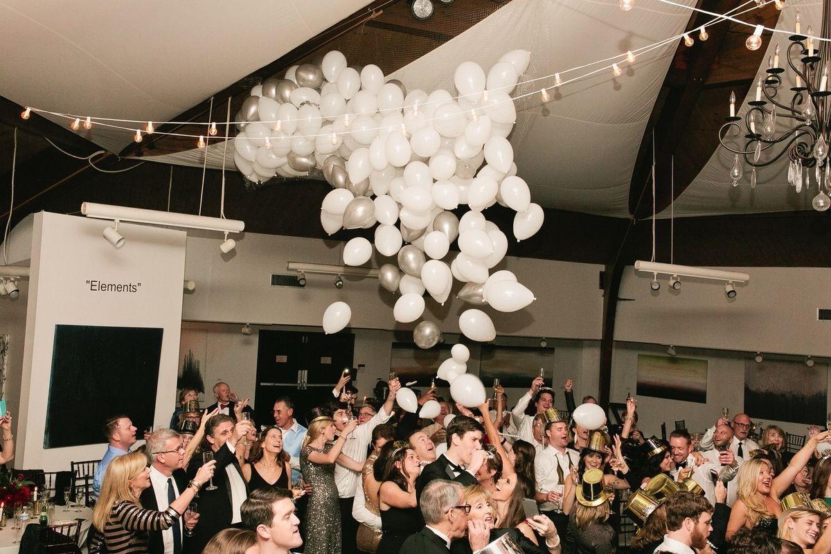 A large cluster of white balloons floats above a celebratory crowd inside a dimly lit event space with string lights.