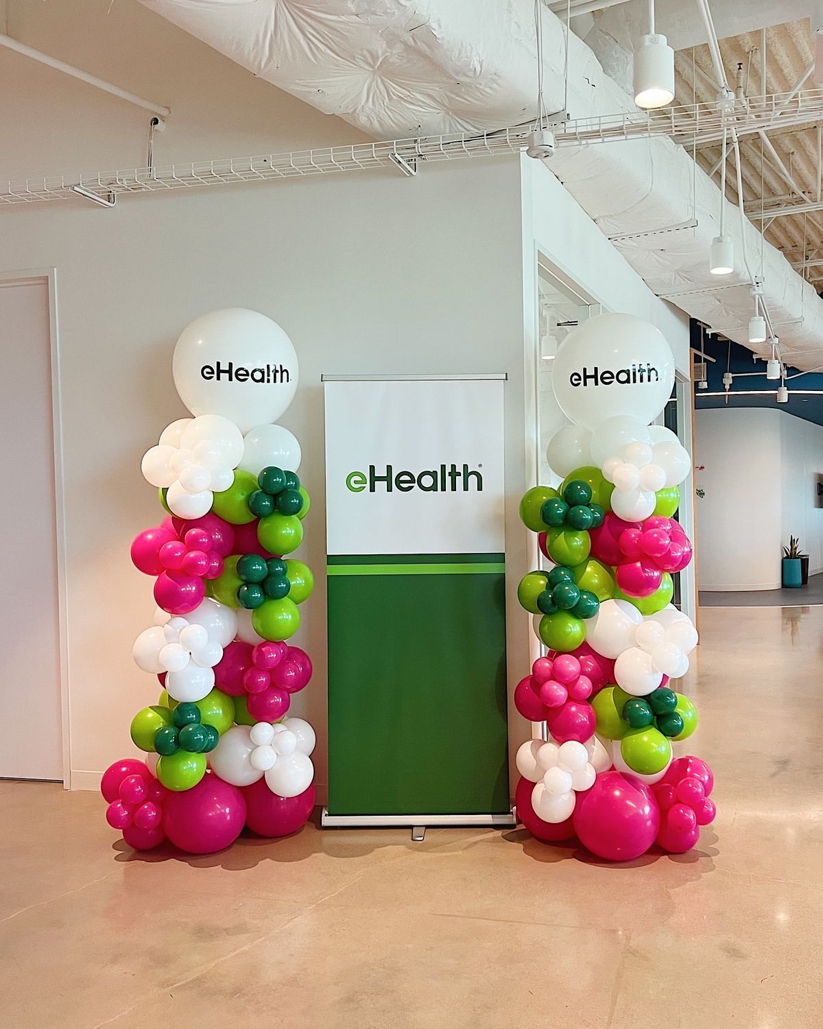 Two balloon columns with white, pink, and green balloons, flanking a banner that says
