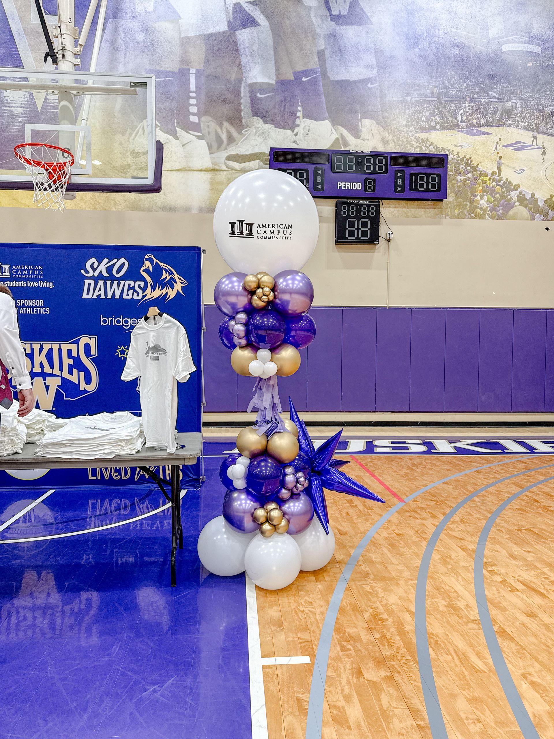 Basketball court with purple and gold balloon decorations; a table with shirts.