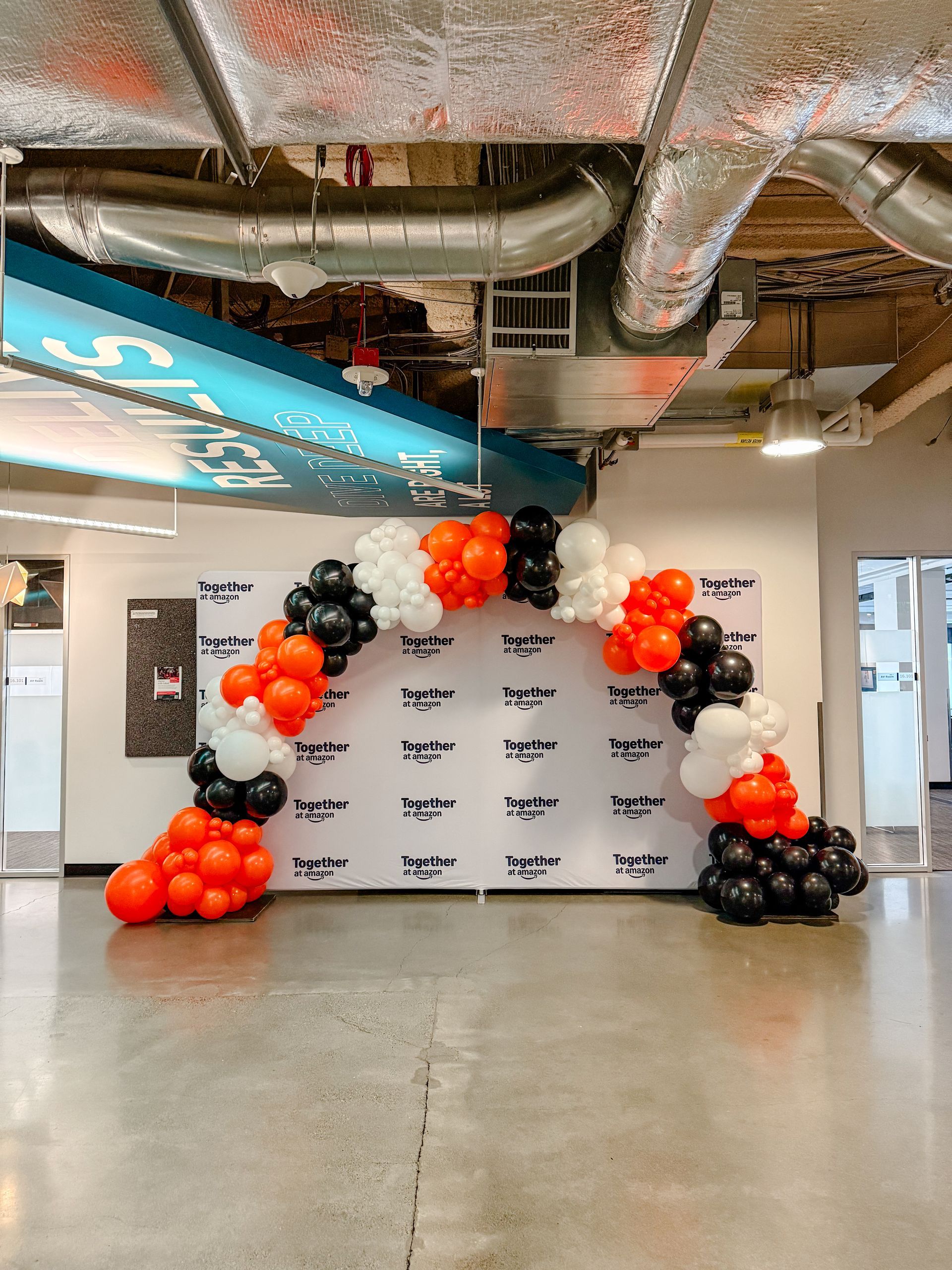 Balloon archway with orange, black, and white balloons in front of a white backdrop with text in a modern office space.