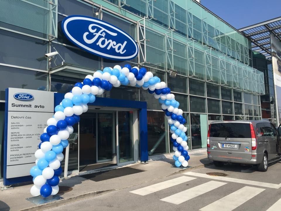 Ford dealership entrance with blue and white balloon arch. A van is parked on the street.