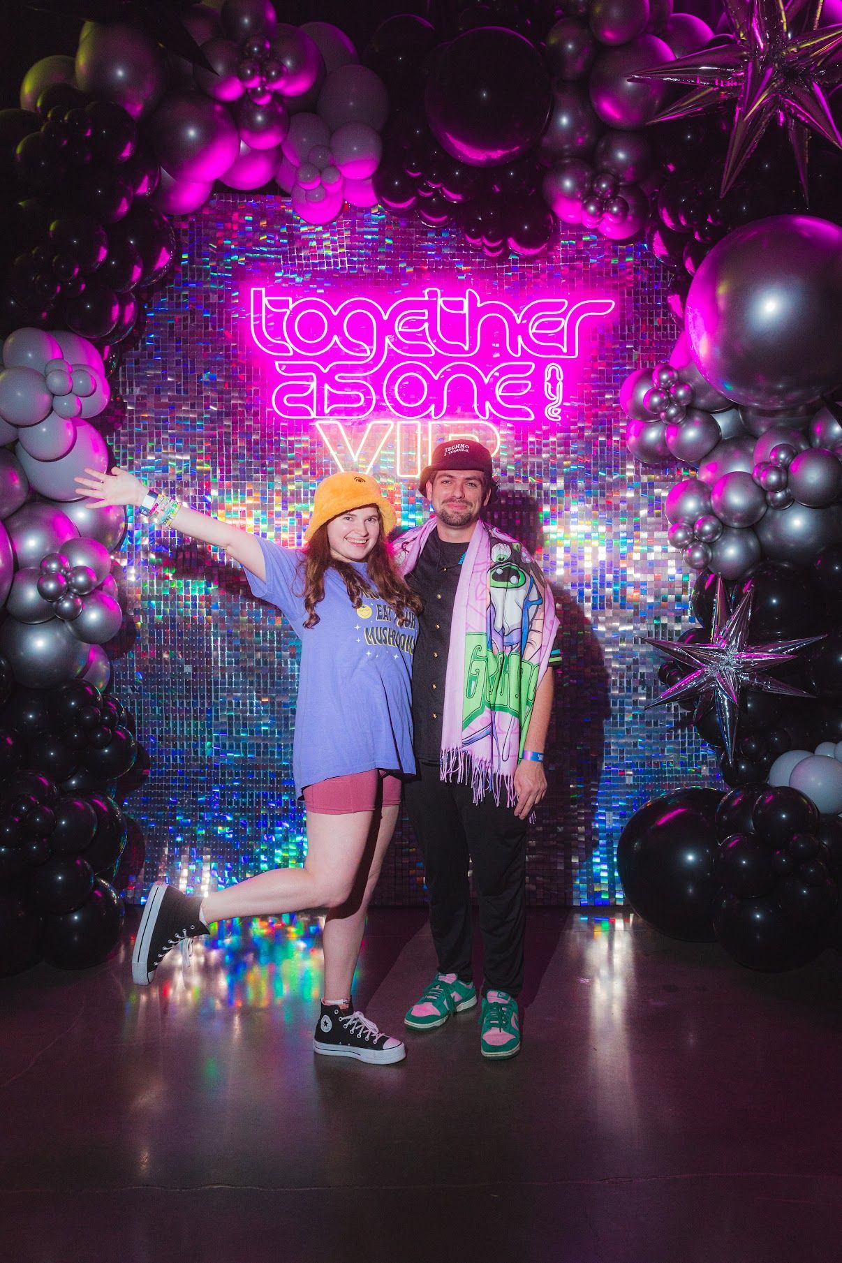 Two people posing in front of a neon sign that reads 
