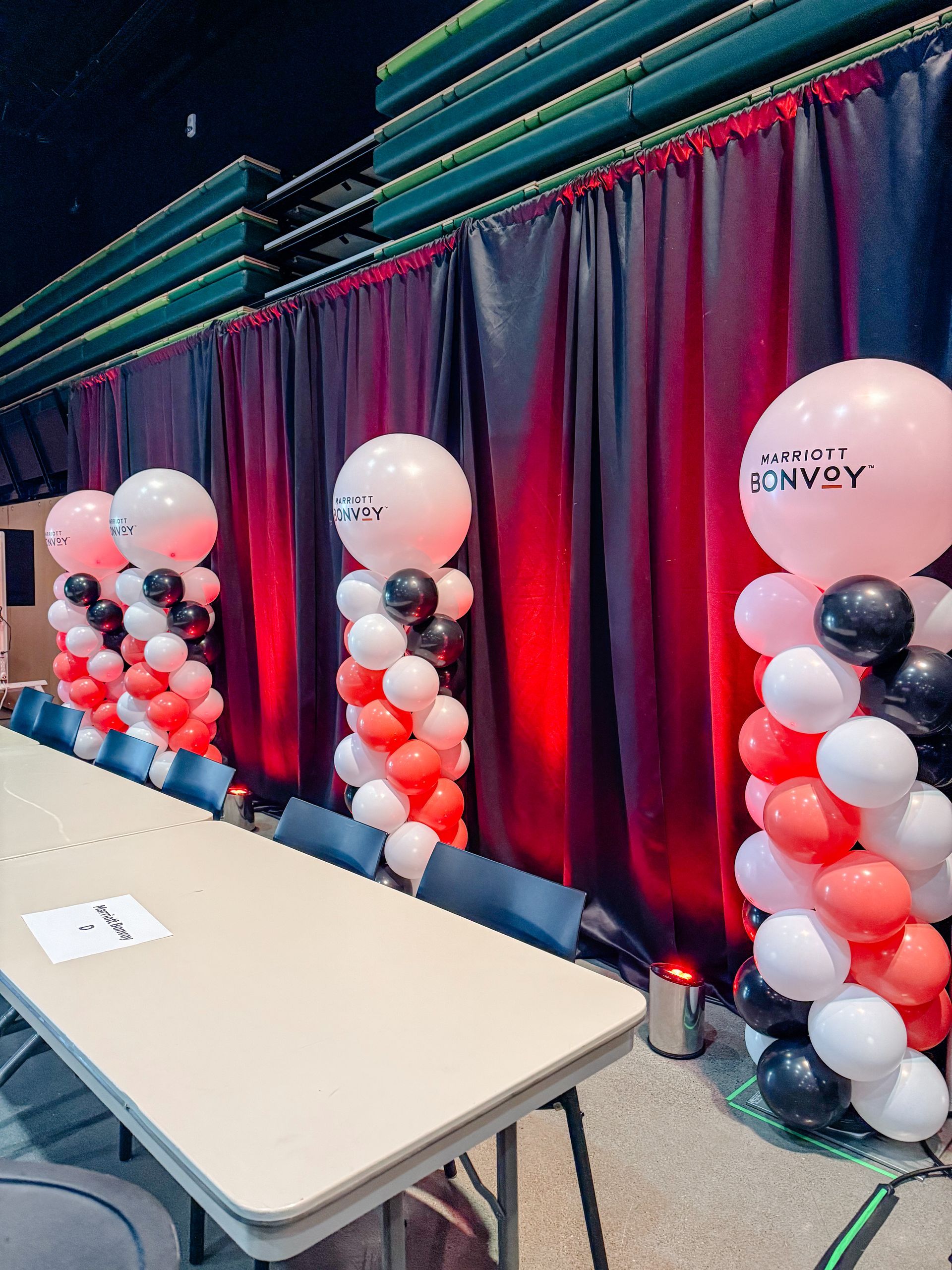 Event setup: Balloons on columns beside a table, red and black curtains in background.