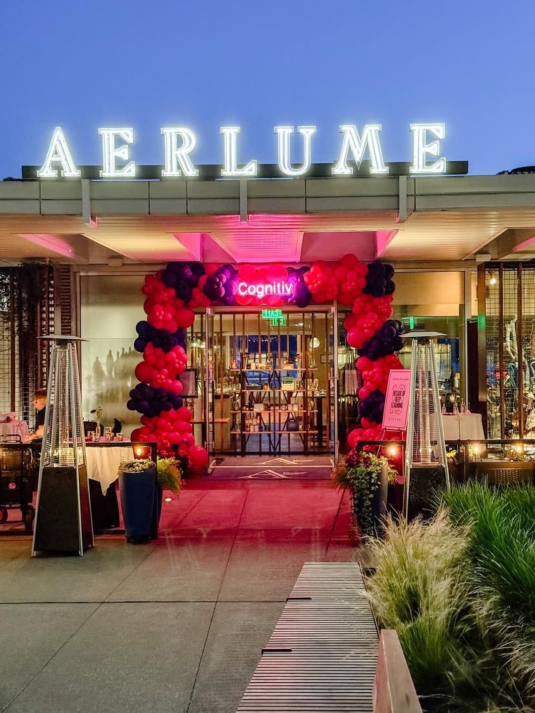 Entrance to Aerlume restaurant with lit-up sign, red and black balloon arch, and outdoor seating.