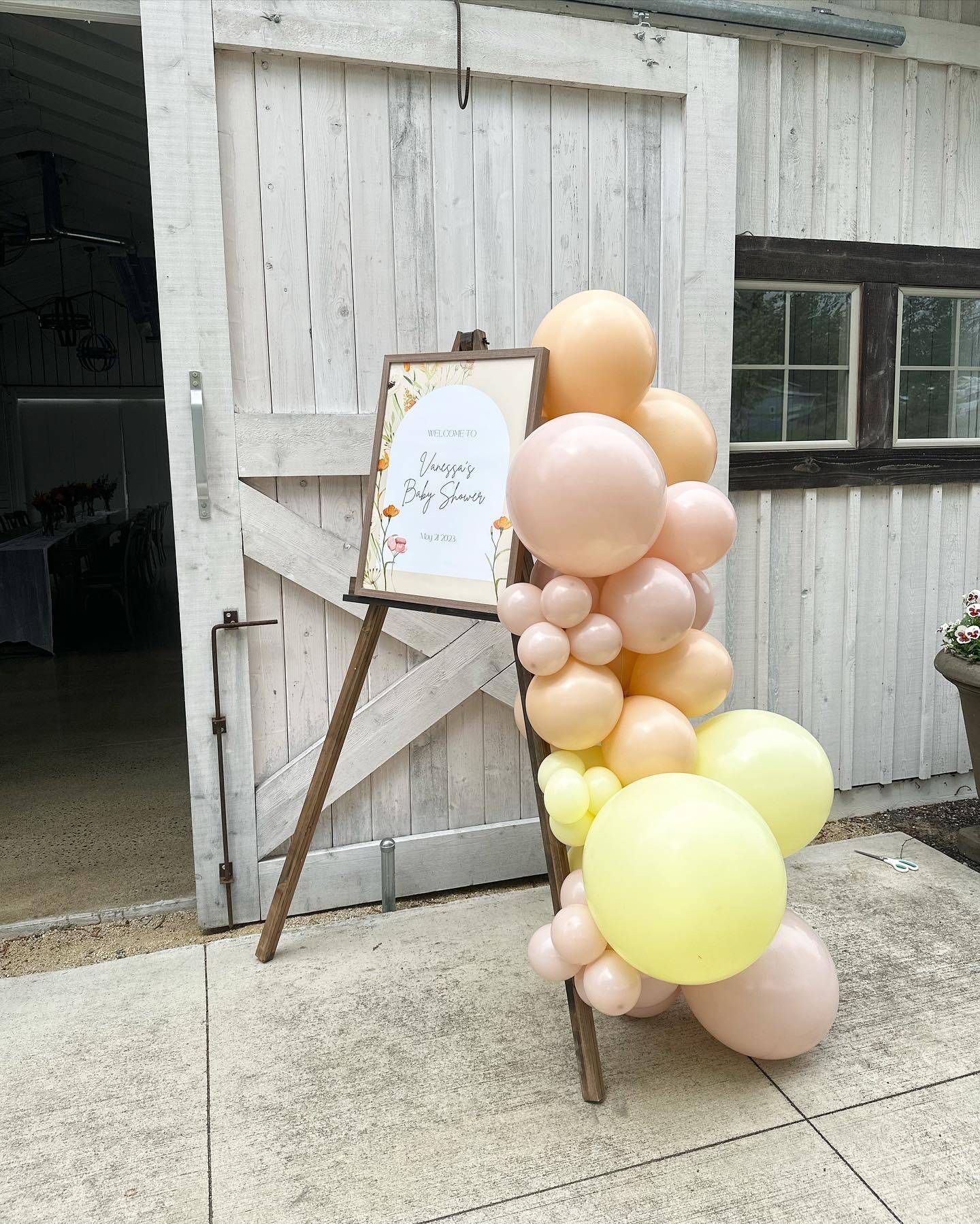 A wooden easel filled with balloons and a sign in front of a barn.