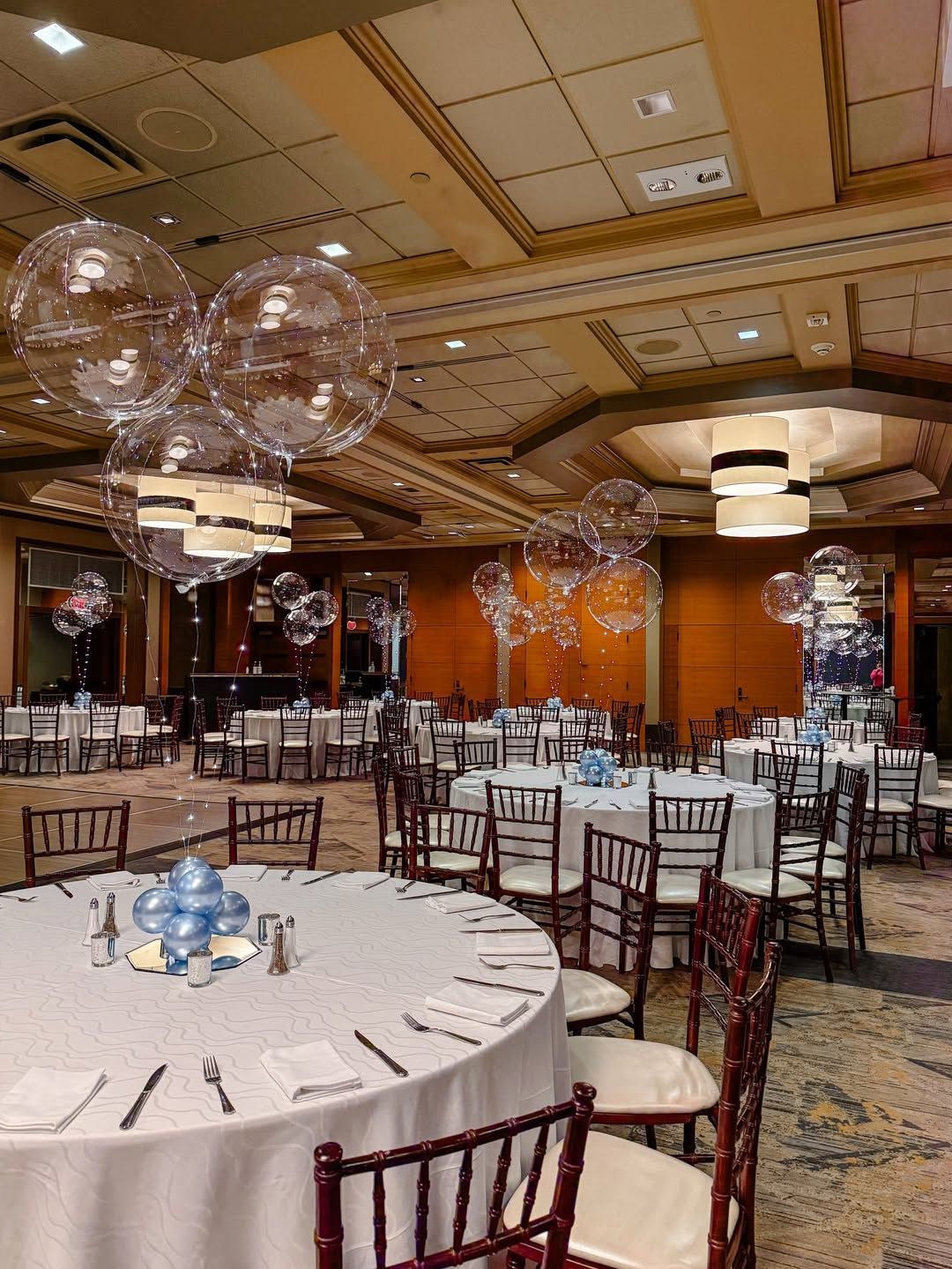 A banquet hall with round tables set for a formal event, featuring clear bubble balloons and blue centerpieces.