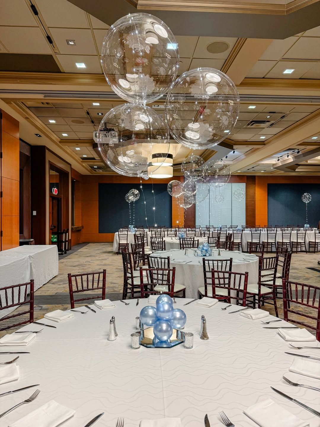 A banquet hall set for a formal event with round tables featuring clear, floating balloon centerpieces and blue accents.