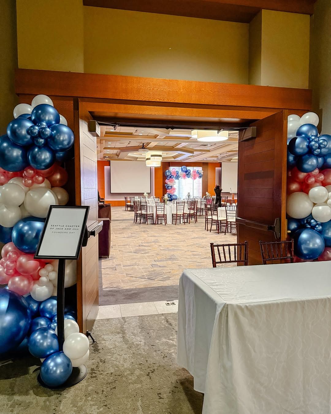 A doorway framed by red, white, and blue balloon arches, leading into a banquet hall with tables and event seating.