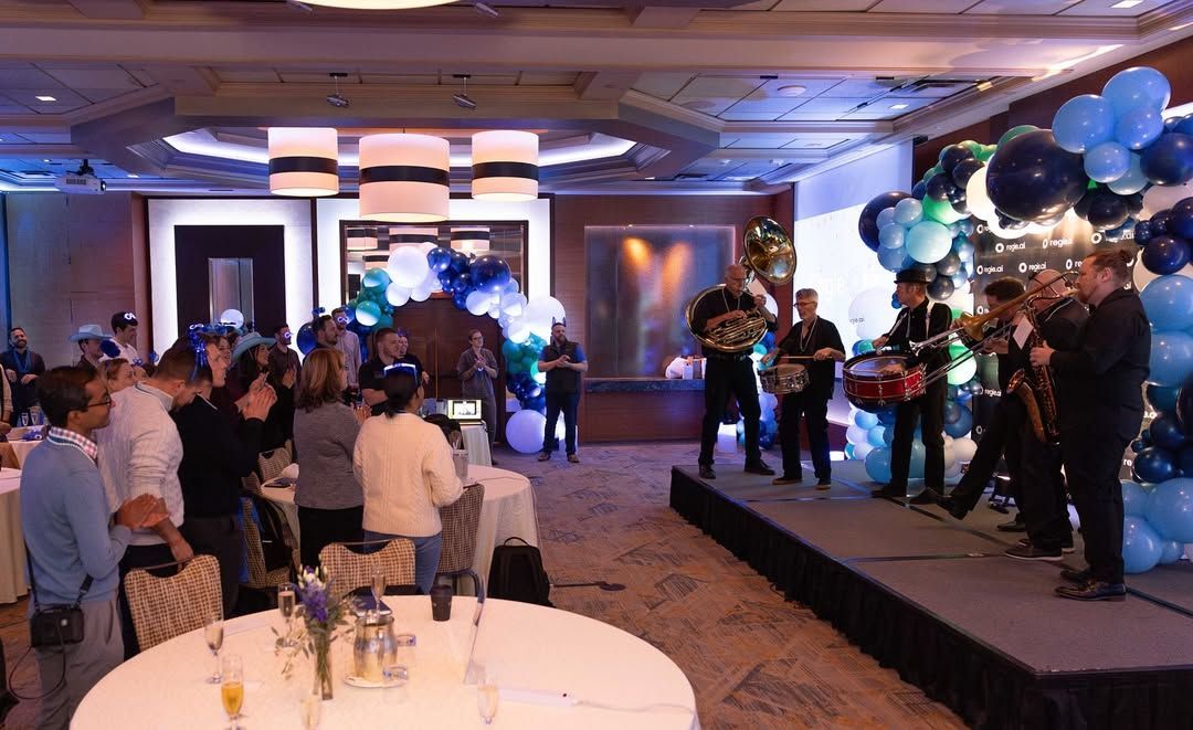 A band performs on a stage for a seated audience in a hotel ballroom decorated with blue and white balloon arches.