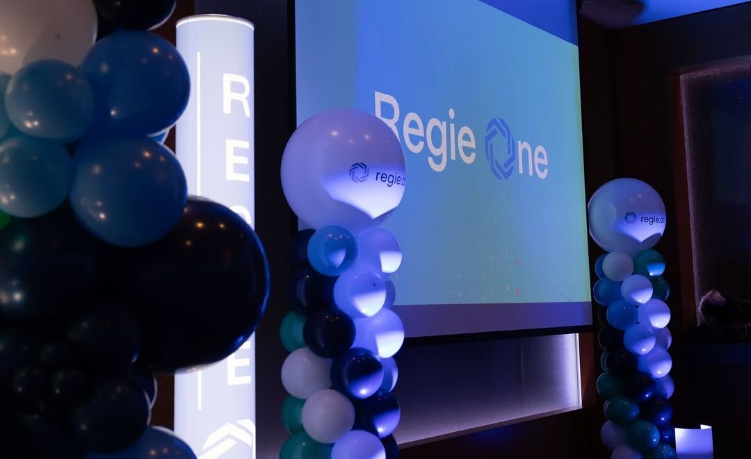Blue and black balloon pillars flank a large projector screen displaying the Regie One logo in a darkened indoor event space.