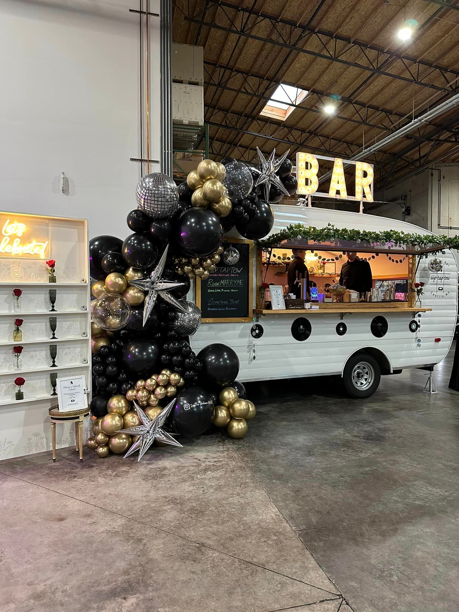 A bar truck is decorated with black and gold balloons in a warehouse.