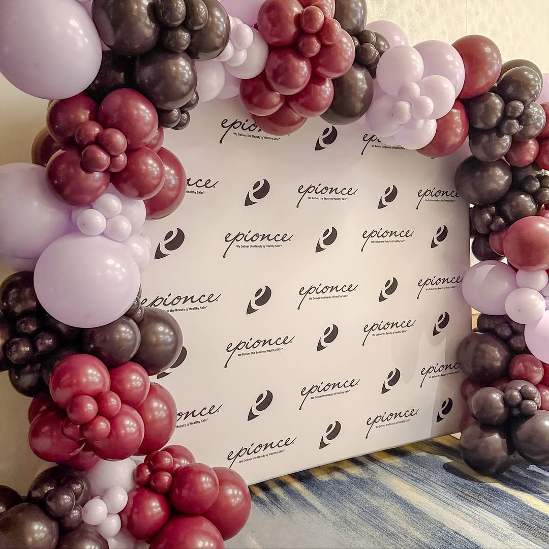 Balloon arch in burgundy, gray, and lavender colors frames a white backdrop with logo.