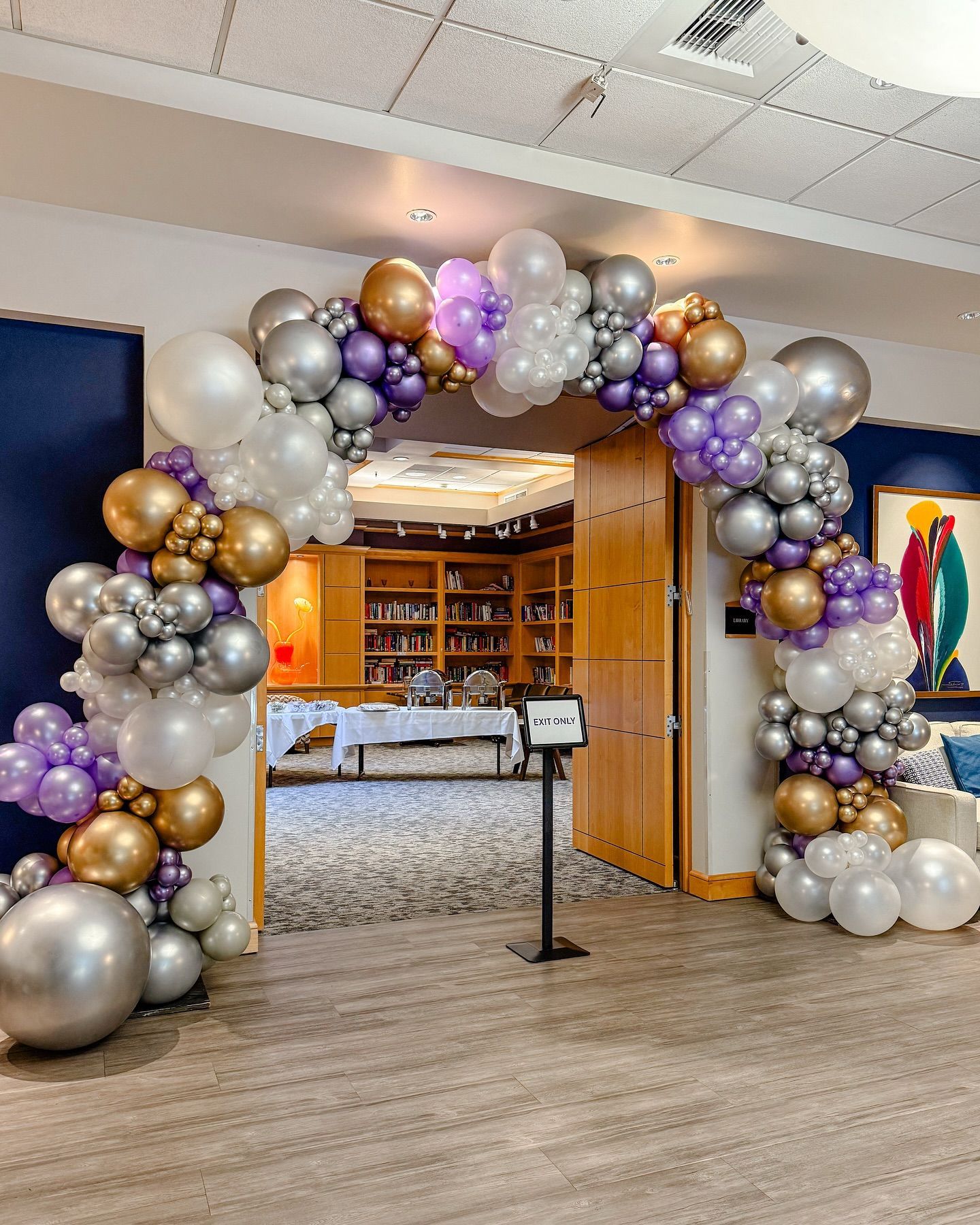 Balloon arch in silver, gold, and purple over a library entrance with open wooden doors.