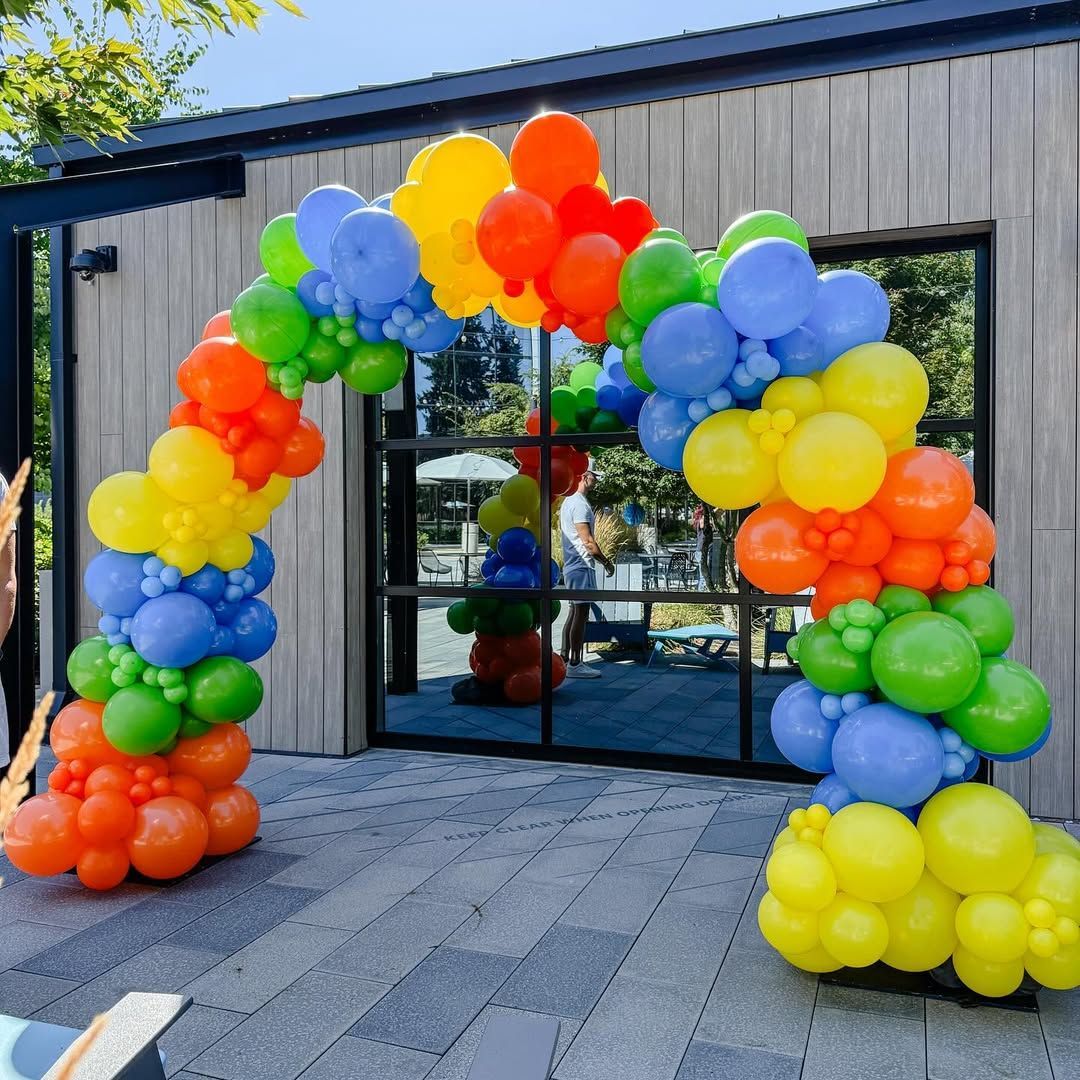 Rainbow balloon arch at a building entrance with glass doors and gray siding